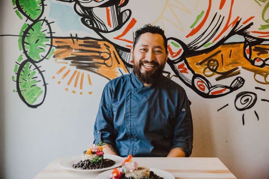 Chef Zaid Consuegra wearing a blue chef's jacket seated in front of a colorful mural with two plates of food on the table in front of him.