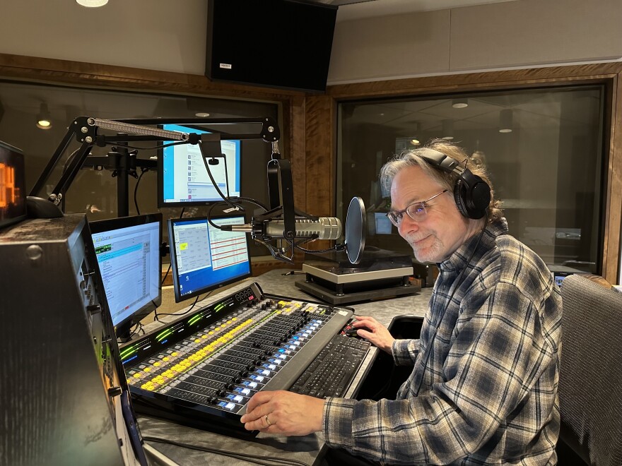 Studio One host Mark Simmet wears a flannel shirt and headphones as he sits at a soundboard with multiple computer screens in front of him.