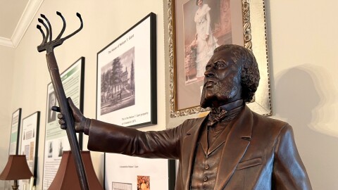 A statue of Nelson T. Gant shows him dressed in a suit and holding a farm tool in his right hand. Behind him, framed posters describe the history of the time.