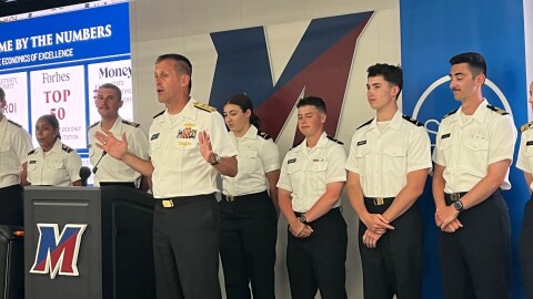 SUNY Maritime College President and retired Rear Adm. John Okon speaks in front of a podium with the letter "M" for SUNY Maritime College. Cadets in uniforms with white shirts and black pants stand in a line behind him. 