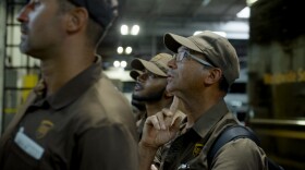 A few UPS workers stand in a warehouse, looking to the left toward something outside the frame of the image.