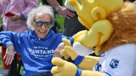 A woman sits outdoors on a sunny day. She is wearing a blue sweatshirt that reads "The City of Fountains Foundation." She is smiling and appears to be talking to a bright yellow mascot lion that is wearing a Kansas City Royals shirt.