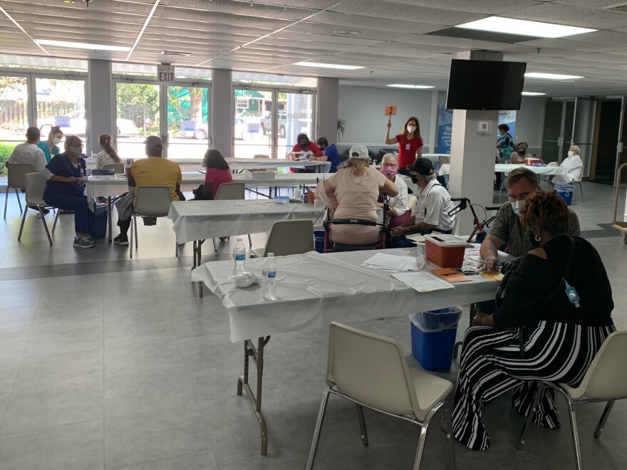 Men and women sitting in front of folding tables in a church community room. Some are health workers administering coronavirus vaccines to seniors of color.