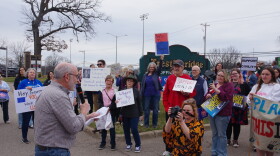 A older man in a checkered white, blue and red button up holds a script as he speaks to a crowd of protesters. The large group hold up signs, some criticize U.S. Representative Bill Huizenga, others call for climate action or criticize the actions of President Donald Trump and Elon Musk.