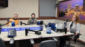 Three people sit at a table in a radio talk studio: a man at left has brown curly hair, a brown beard and is wearing glasses, a mustard yellow button-down shirt and white t-shirt; a man at center has a dark goatee and is wearing a black knit cap and green sweatshirt; a woman at right has long red hair and is wearing a black and white striped button-down shirt.