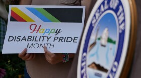 Hands hold a sign that reads "Happy Disability Pride Month" in the background. In the foreground, the seal of the city of Buffalo is blurred.