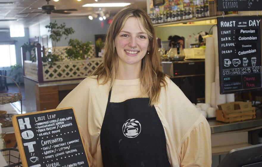 Woman in a tan top and black apron standing behind the counter at a coffee shop and next to a sign listing a coffee menu.