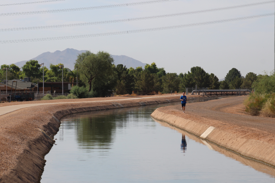 A person runs along a dusty trail beside a canal. Trees and a couple houses are visible in the background.