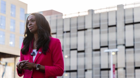 A woman in a pink blazer and a small microphone clipped to her lapel stares ahead.