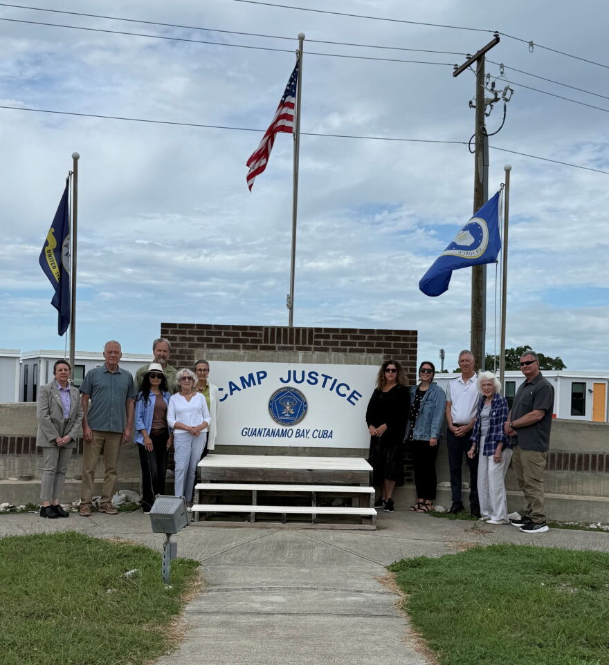A group of people is standing near a brick sign that says Camp Justice, Guantanamo Bay, Cuba.