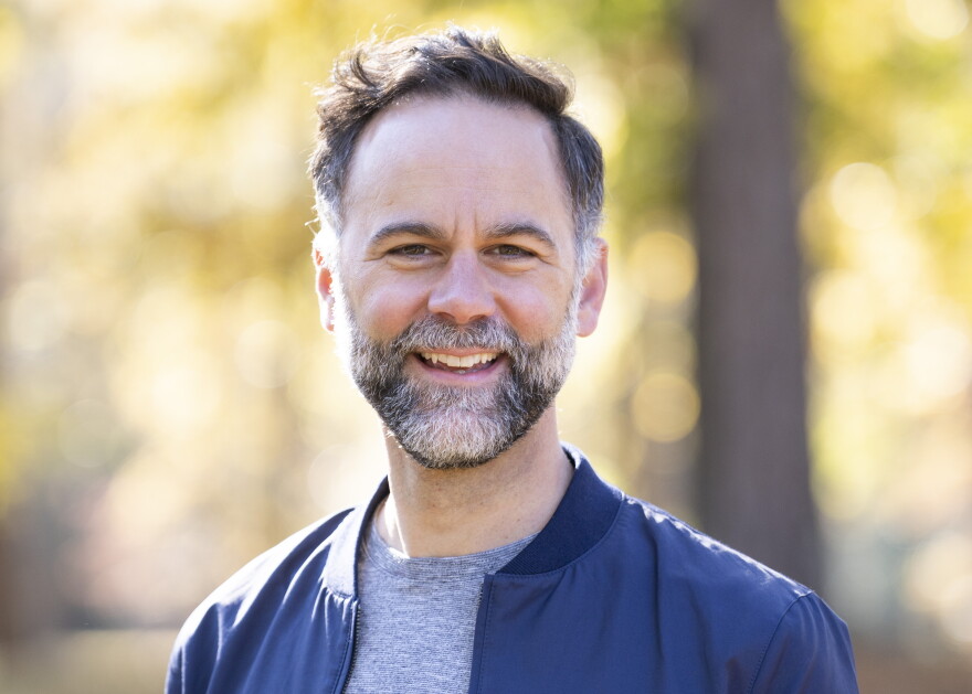 Smiling man with beard wearing a blue jacket with bright, blurred foliage in the background