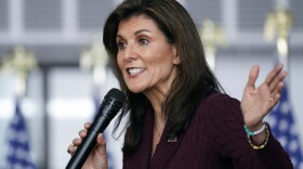 A woman holds a microphone and gestures while speaking in front of a line of flags