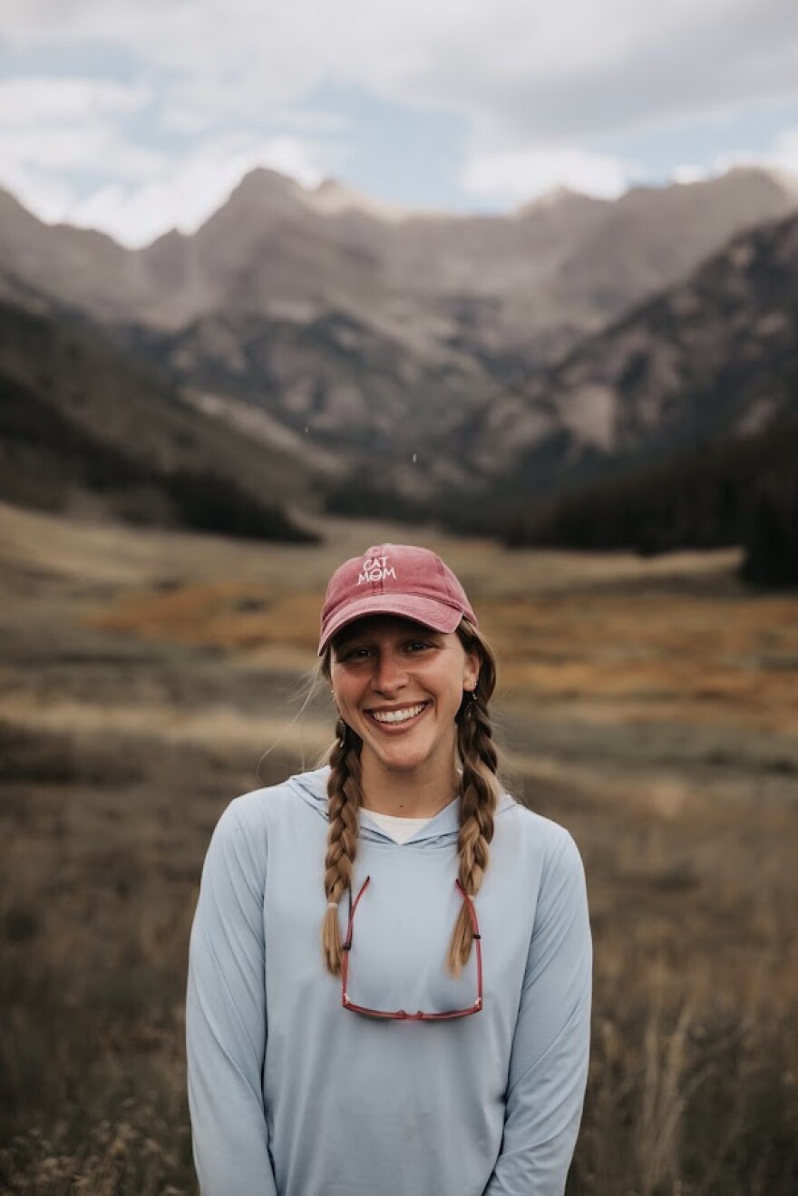 A woman wearing a light blue hoodie and a faded red ball cap, with her hair in two long, light-brown braids, smiles for a photo with mountains in the background