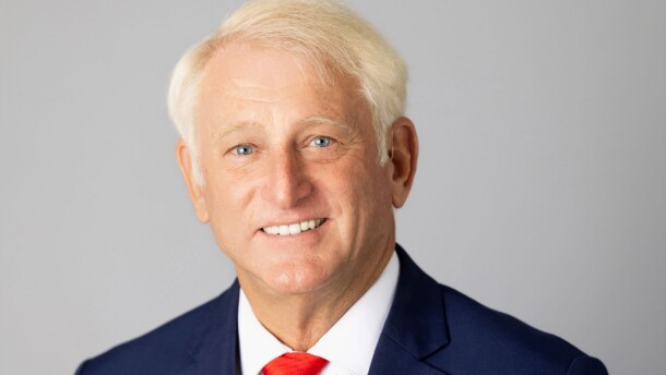 A headshot of a man with white hair, blue eyes, a blue shirt and a red tie