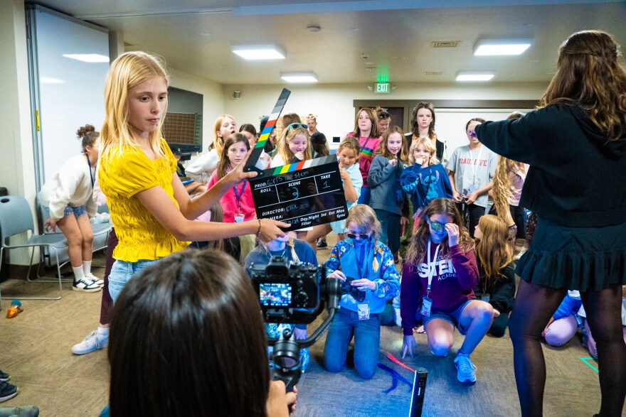 A middle school girl in a yellow shirt holds up a clapperboard in front of a black camera. Behind her is a large group of young students. They're inside a classroom with carpet flooring and whiteboards on the walls. 