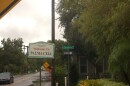 A vintage sign reads "Welcome to PALMA CEIA" on a street corner. Lush greenery, one house and one car are visible in the background.