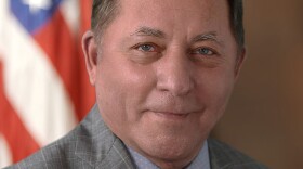 A smiling man with short grey hair wearing a grey blazer with a checked pattern and a pin, a blue collared shirt, and a blue tie with a white pattern. He is sitting in front of an American flag. 