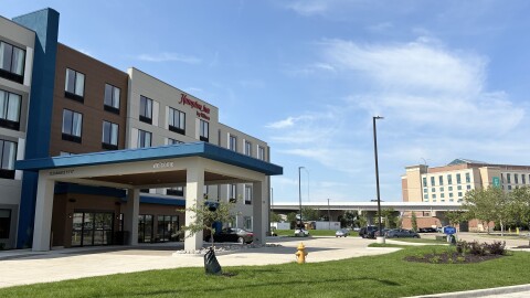 A Hampton Inn hotel building with a covered entrance and a "welcome" sign, surrounded by green grass, trees, a fire hydrant, and a few parked cars, with another hotel visible in the background under a blue sky.
