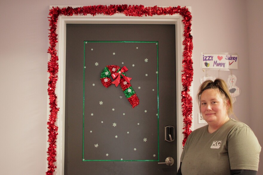 A woman stands beside a door decorated with a candy cane and red and green decorations for Christmas.