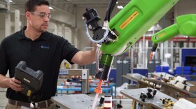 An employee is working alongside a FANUC "cobot" that is applying adhesive to a glass windshield.