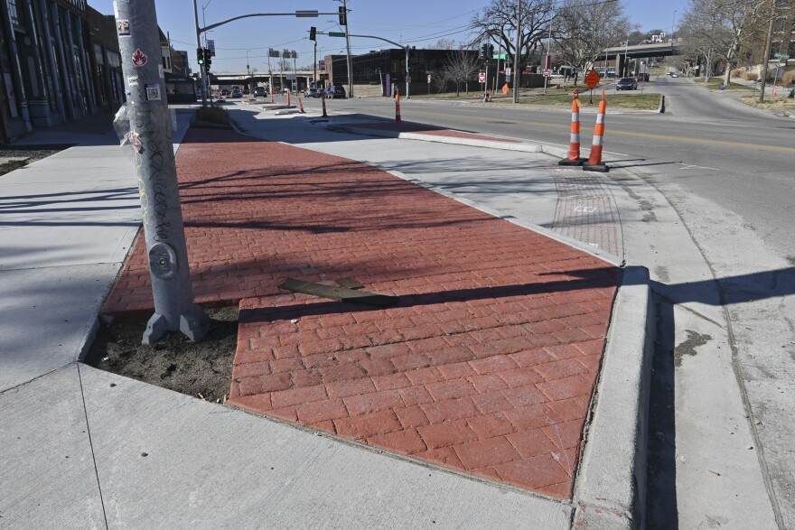 Exterior photo of a sidewalk near an intersection. In foreground is a red brick inset near some freshly poured concrete. In background are traffic cones