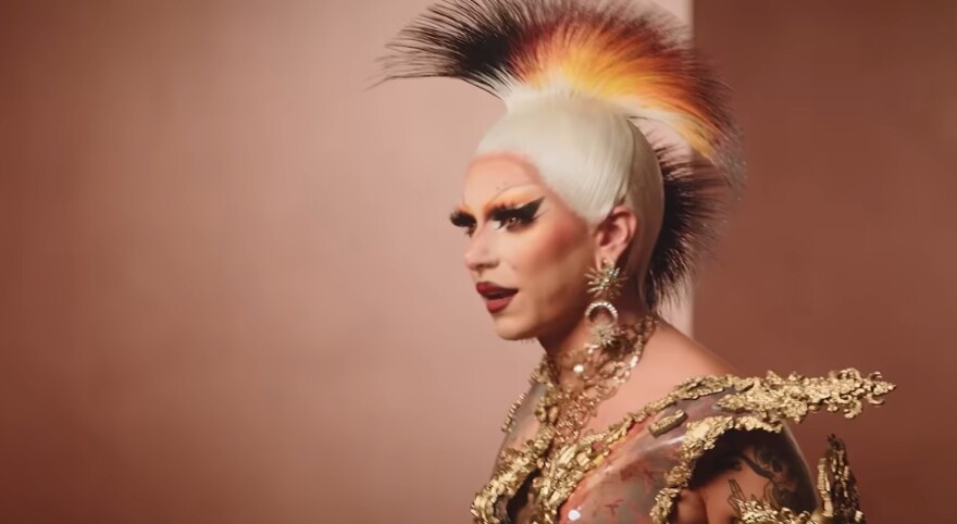 A drag queen looking left with a gold and black mohawks and gold outfit