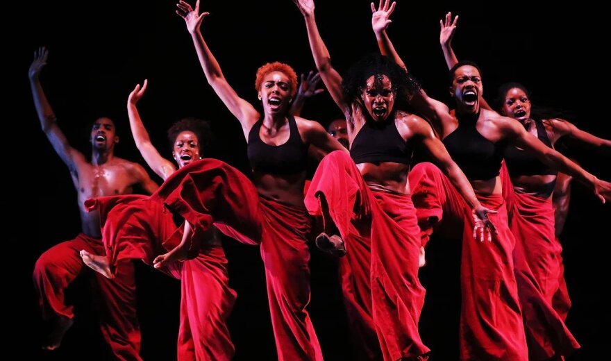 Several dancers wearing flowing red pants and black tank tops dance together in a performance with their arms raised.