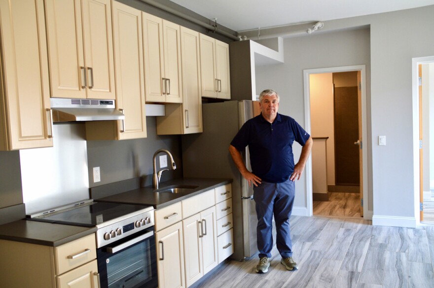 A man in jeans and a blue T-shirt stands in front of a modern kitchen he's added to a newly renovated downtown Rutland apartment.