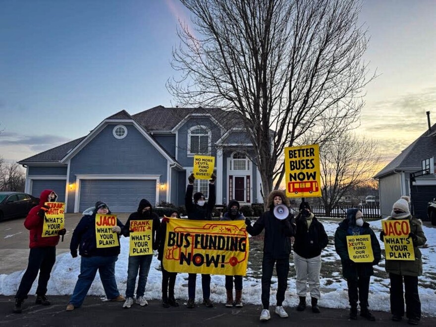 A group of people holding yellow signs stand in front of a blue house