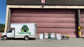 A white truck that says "Alaska Plastic Recovery Home of the Grizzly Wood" is parked in front of an oversized garage with a red door. Two big white plastic bags and two people in camping chairs are next to the truck.