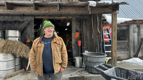 Man in green hat and work coat standing in front of his sugarhouse 