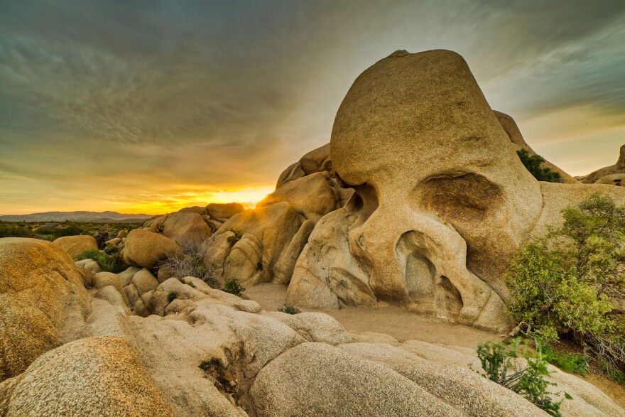 A skull-like rock formation in a desert landscape at sunset, with golden light illuminating distinctive granite boulders against a dramatic sky with orange and blue hues.