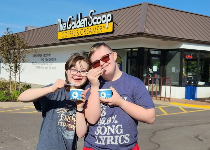 A woman and man stand together posing for a photo in front of The Golden Scoop Coffee and Creamery. Each person has a cup and spoon in their hand. 