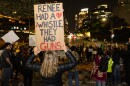 A woman holds a sign reading "Renee had a whistle, they had guns" above her head. She is in the foreground and a crowd and downtown street scene is seen in the background. 