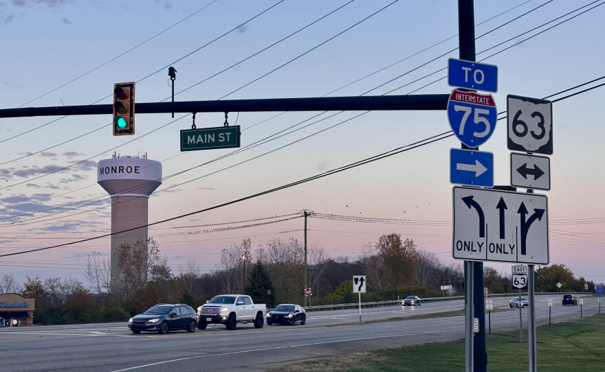 Cars sit stopped at a red light with a sunset and Monroe water tower in the background.