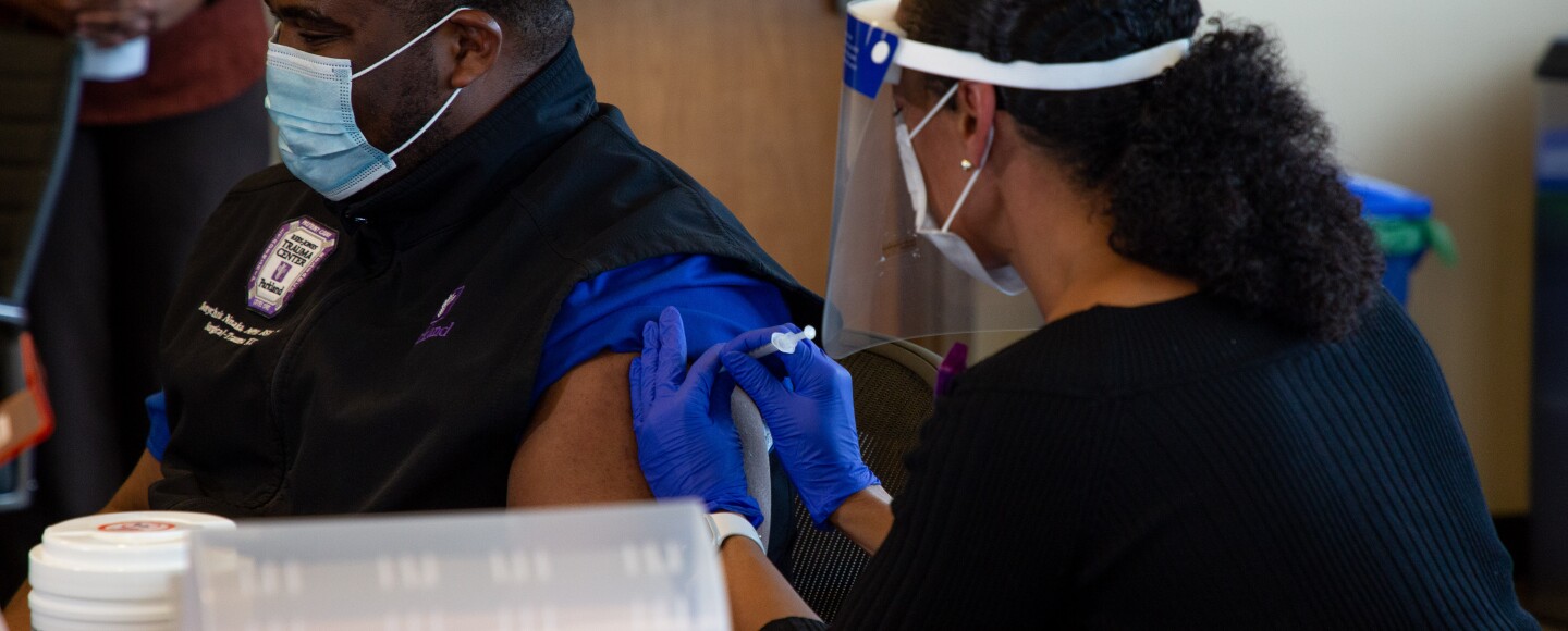 A nurse wearing gloves and a face shield administers a shot to a nurse who works on the COVID-19 team at Parkland Hospital.