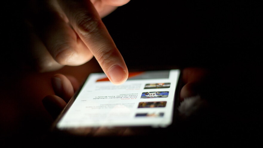 A person uses their index finger to scroll through a list of news articles on their smartphone. The screen lights up the centre of the photo as the surrounding background is low light.