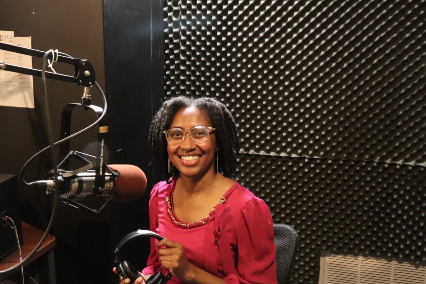 A smiling woman in a pink blouse sits in a recording studio holding headphones beside a microphone, with soundproof foam panels behind her.