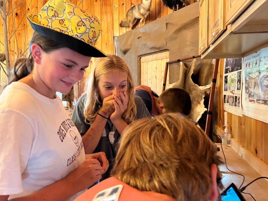 A girl in a hat with yellow rubber ducks peers at another boy, who is looking into a microscope. Beside her, a girl covers her mouth with her hands in fright.