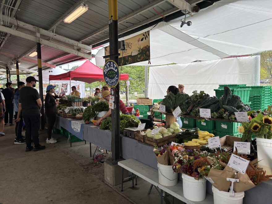 A farm stand displays flowers and vegetables, while a worker moves produce and shoppers browse items