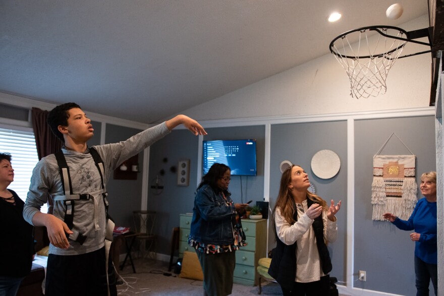 At their home in Jenks, Kannon Cooper threw a ball toward a basketball net mounted in his living room while his sister, Kellee Cooper, waited to catch it on March 14, 2023 The goal is one of many functions in the family's home designed to accommodate Kannon who is 14 but thinks and acts like a toddler due to a rare condition called Lennox Gastaut Syndrome. On her phone in the background is Kannon's case manager, Cheryl Dever, who works for the state's Development Disability Services. Dever is the family's fourth case manager since being approved for services in October.