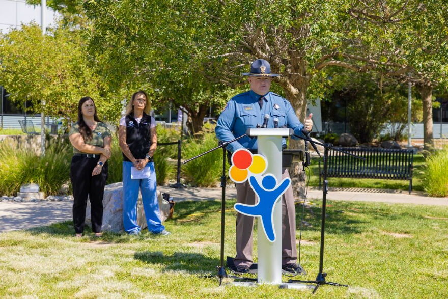 A man in a state trooper uniform stands at a podium, addressing an audience. Two women stand behind him, looking on.