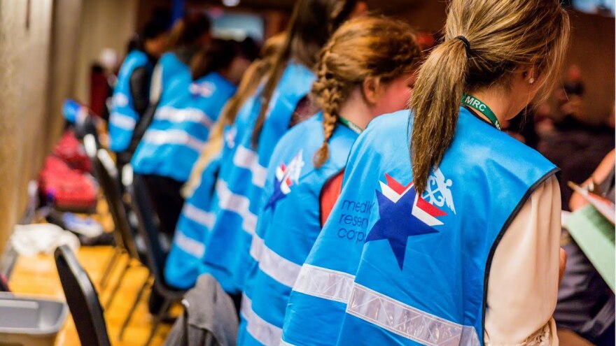 Photo of a number of people in a line. Visible is the back of their blue vests that reads Medical Reserve Corps. 