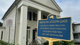 A white Greek Revival style home in the background with an historic marker sign in the foreground. The sign says, "MATILDA JOSLYN GAGE: Nationally known abolition and women's rights advocate lived here from 1854 until her death in 1898."