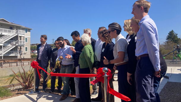 A person wearing a button-down shirt and sunglasses cuts a red ribbon alongside several other people.