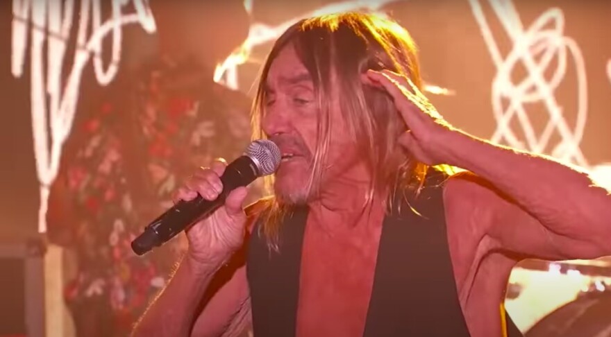 Iggy Pop is singing into a microphone while wearing a black vest.