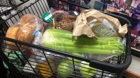 A shopping cart is filled with groceries, including celery, bread, green apples, and canned goods.