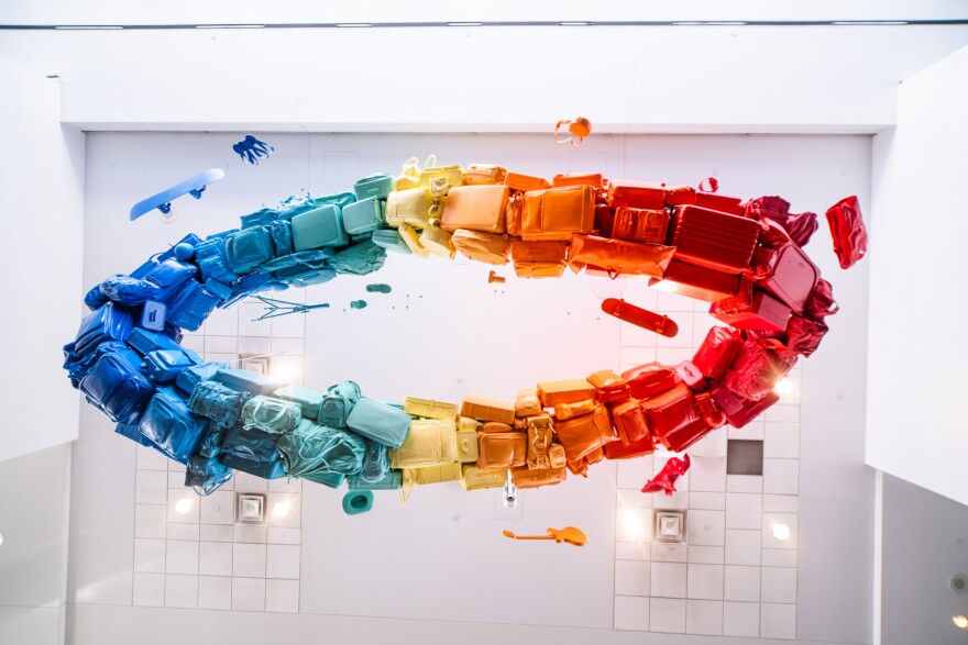 Painted bags of luggage in a circle with a rainbow of colors hang from an all white ceiling.