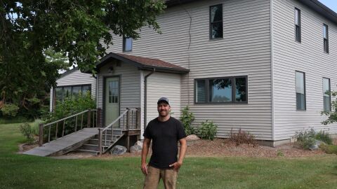 A man in his forties wearing a black shirt stands in front of his home, which has stood for over a hundred years.