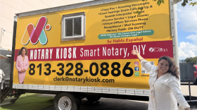 Una mujer con un vestido blanco posa frente a un tráiler que tiene en letras blancas en el frente la leyenda Notary Kiosk Smart Notary DIY.